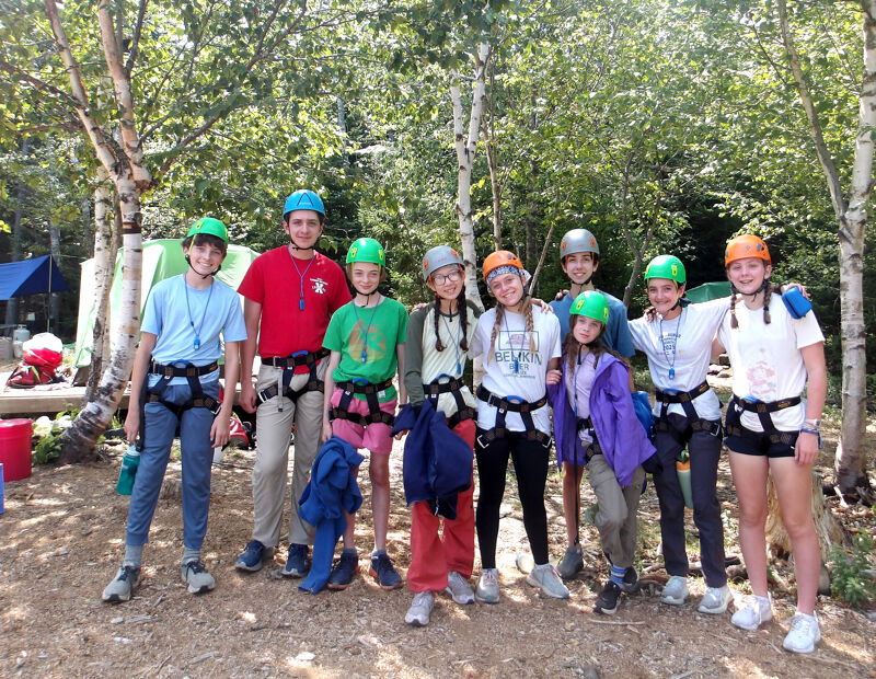 The image shows a group of young people wearing helmets and harnesses, likely at a ropes course or adventure park. They are standing outdoors in a wooded area, with trees and a tent visible in the background. The group appears to be posing for a photo, with smiles on their faces, suggesting they are enjoying an activity.